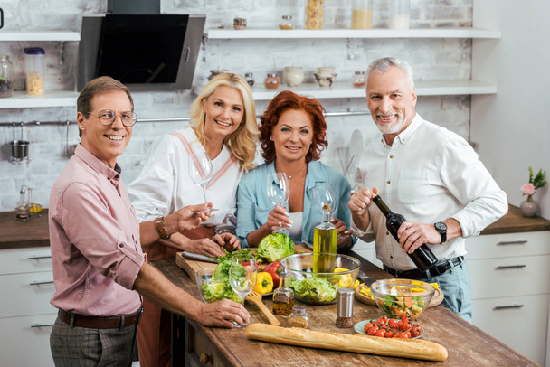 mature friends celebrating meeting with salad and wine in kitchen, looking at camera - Photo, Image