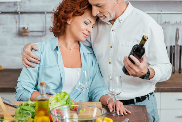 affectionate husband hugging wife during salad preparation for dinner in kitchen and holding wine bottle - Фото, зображення
