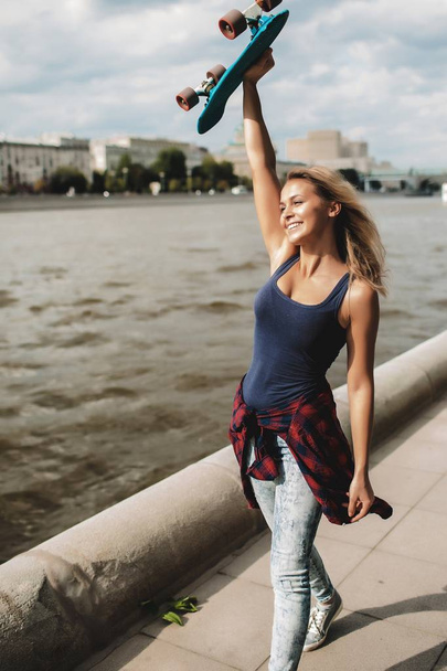 happy blonde woman holding skateboard above head and walking at river  - Фото, изображение