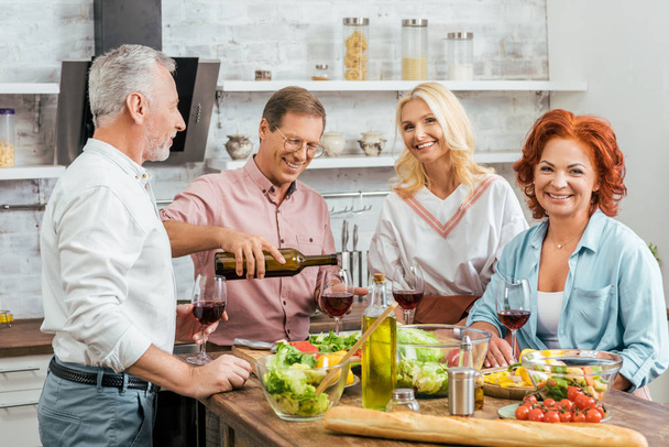 handsome man pouring wine to happy old friends during dinner in kitchen - Photo, Image