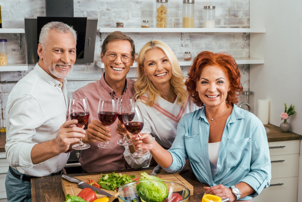 smiling old friends clinking with wineglasses in kitchen and looking at camera - Photo, Image