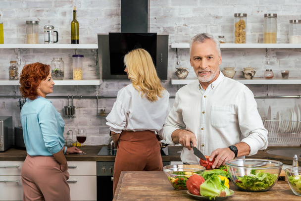 handsome man preparing salad for dinner at home, women talking with wine - Photo, image