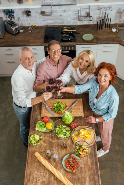 high angle view of smiling old friends clinking with wineglasses in kitchen - Photo, Image