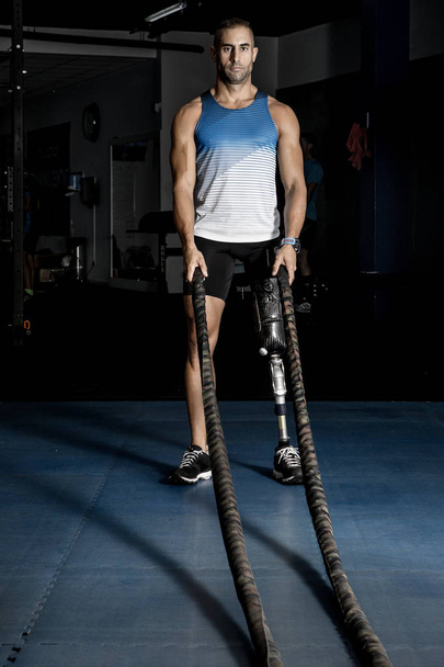 Erwachsener Athlet mit Beinprothese steht mit Kampfseilen in Turnhalle - Foto, Bild