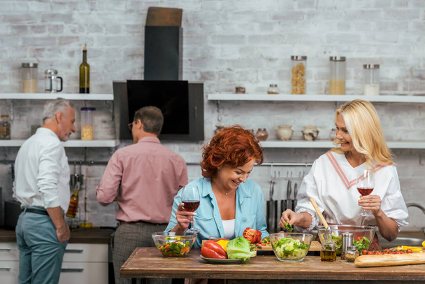 laughing beautiful women preparing salad for dinner and holding wineglasses at home - Photo, Image