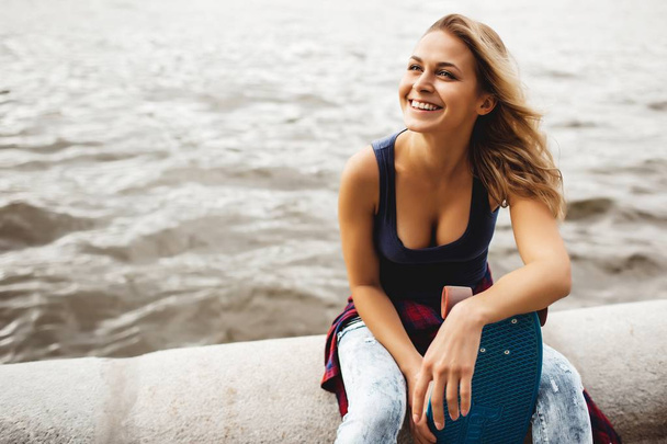 smiling blonde teenager girl with blue skateboard sitting at river on road border - Foto, Bild