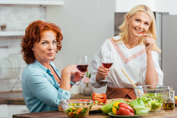 smiling attractive women preparing salad for dinner, holding wineglasses and looking at camera at home - Foto, immagini