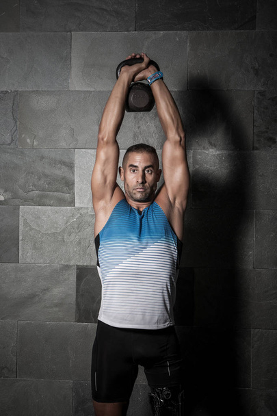 Adult confident man with prosthesis lifting heavy kettlebell above head against gray wall  - Foto, afbeelding