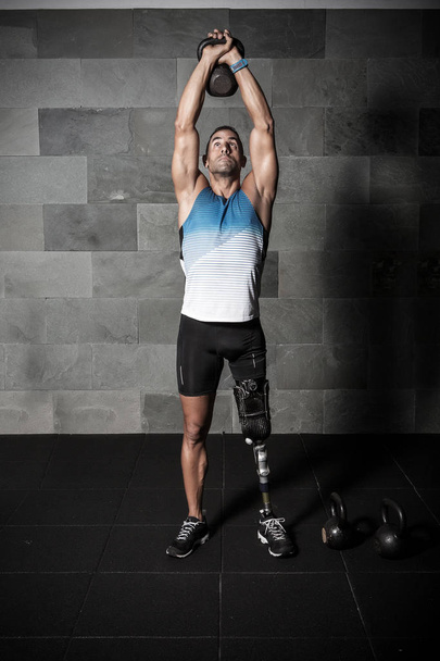 Мускулистый взрослый спортсмен с искусственной конечностью ноги поднимает руки над головой с гирей
 - Фото, изображение