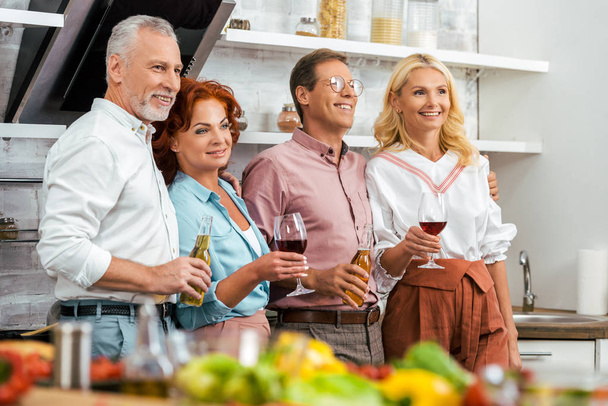 happy mature friends holding alcoholic beverages and looking away in kitchen - Photo, Image