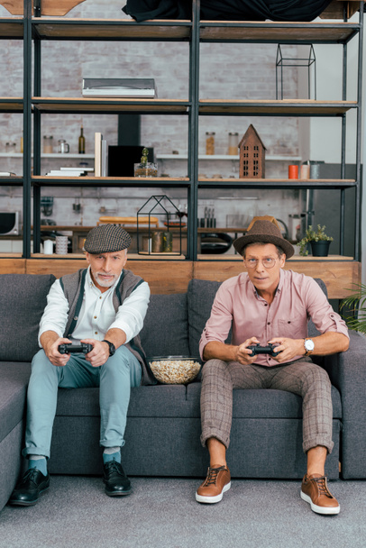 maduros amigos do sexo masculino usando joysticks e olhando para a câmera enquanto sentados juntos no sofá
  - Foto, Imagem