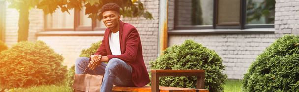 wide shot of smiling young man with leather backpack sitting on bench on street - Photo, Image