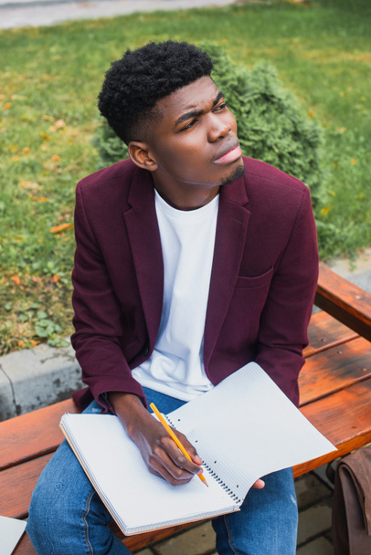 thoughtful young man sitting on bench and writing in blank notebook - Photo, Image