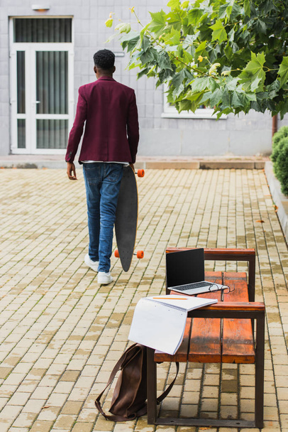 vista trasera del hombre caminando con monopatín en la calle con ordenador portátil y portátil en el banco en primer plano
 - Foto, imagen