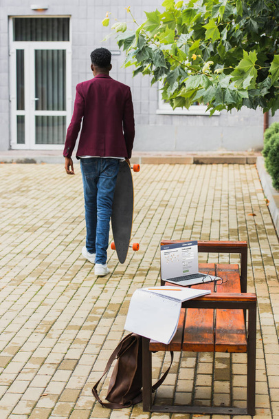 rear view of freelancer walking with skateboard on street with laptop with facebook website on screen lying on bench on foreground - 写真・画像