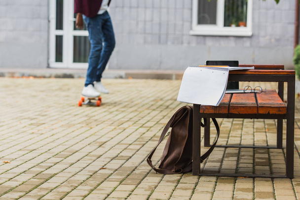 cropped shot of student riding skateboard with backpack and notebook on bench on foreground - Foto, Bild