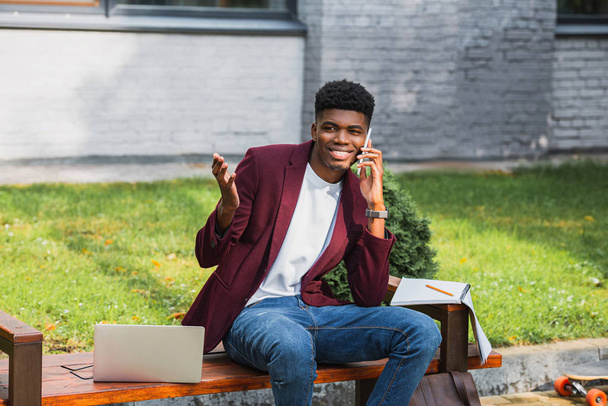 usměvavé mladé freelancer mluví po telefonu na lavici s notebookem - Fotografie, Obrázek