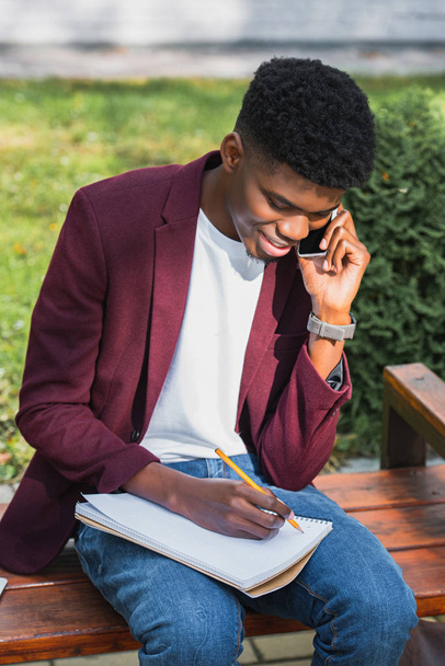 smiling young student talking by phone and writing in notebook sitting on bench - Photo, image
