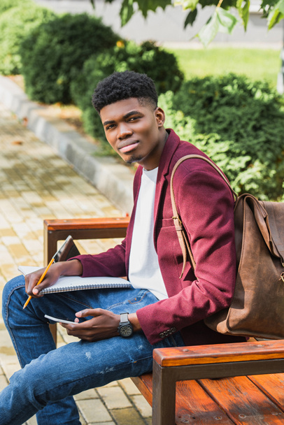handsome young student using smartphone while sitting on bench - Foto, afbeelding