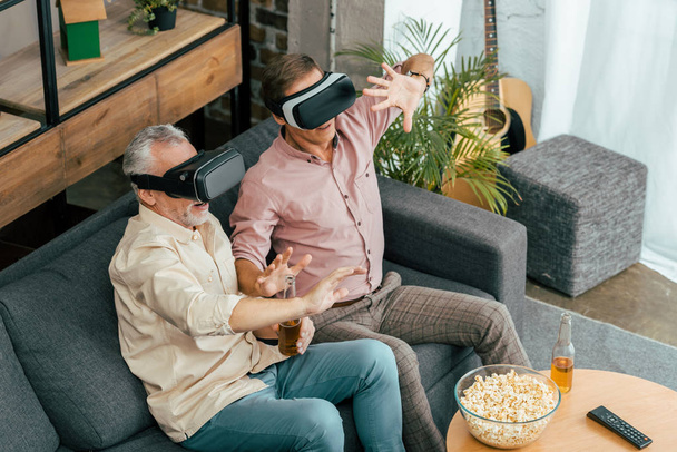hoge hoekmening van volwassen mannen bier drinken en het gebruik van virtuele realiteit headsets thuis  - Foto, afbeelding