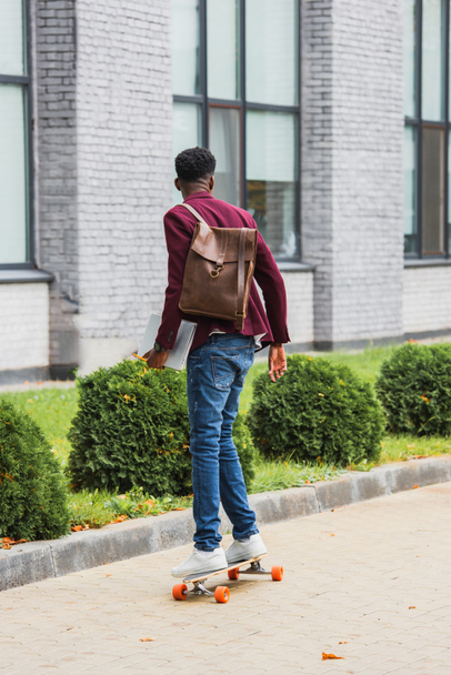 vue arrière de l'élève avec sac à dos et cahiers chevauchant sur planche à roulettes sur rue
 - Photo, image