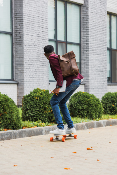 rear view of young student with backpack and notebooks riding on skateboard on street - Фото, зображення