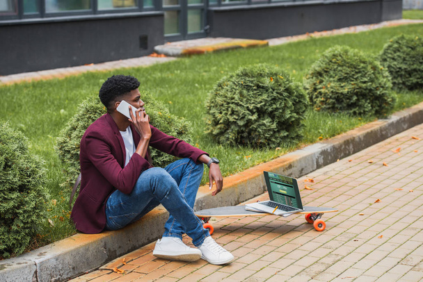 nuori freelancer puhuu puhelimitse istuessaan hillitä kadulla kannettava tietokone urheilun verkkosivuilla ruudulla makaa rullalauta
 - Valokuva, kuva