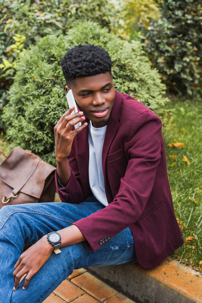 smiling young man talking by phone while sitting on curb on street - Photo, Image