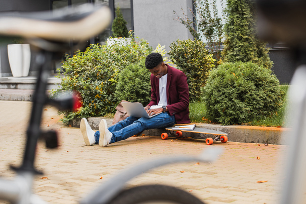 young student using laptop and writing in notebook while sitting on curb on street - Foto, Bild