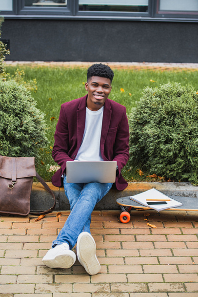 heureux jeune étudiant en utilisant un ordinateur portable tout en étant assis sur le trottoir sur la rue et en regardant la caméra
 - Photo, image