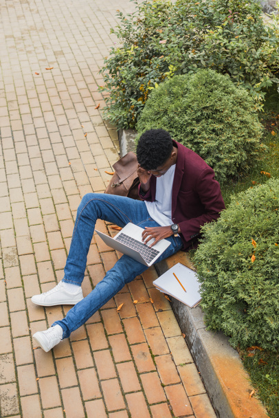 mladí na volné noze práce s notebookem a mluví po telefonu při posezení na chodníku na ulici - Fotografie, Obrázek