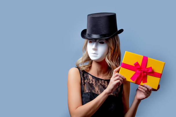 Portrait of a young style girl in top hat and white mask with gift box on gray background - Zdjęcie, obraz