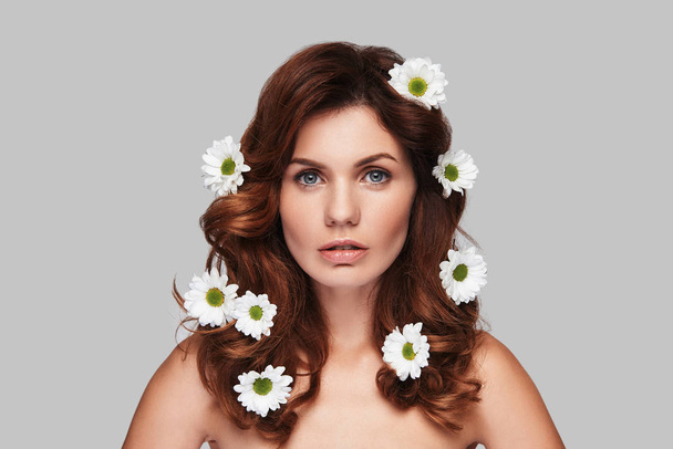 beautiful Caucasian woman looking at camera while standing against grey background with chamomile flowers in hair  - Zdjęcie, obraz