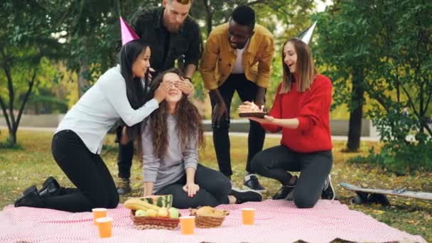 Молодые люди поздравляют девушку с днем рождения, приносят торт обнимает и целует ее, женщина задувает свечи, смеется и радуется во время вечеринки на открытом воздухе в парке
. - Кадры, видео