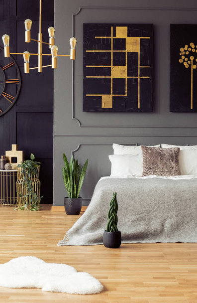 Gold and black painting hanging above bed with bright sheets in real photo of dark bedroom interior with fresh plants and molding on wall - Zdjęcie, obraz