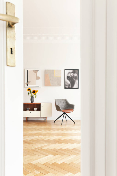 Real photo through the door of bright living room interior with grey chair, cupboard with sunflowers, three posters and herringbone parquet - Фото, зображення