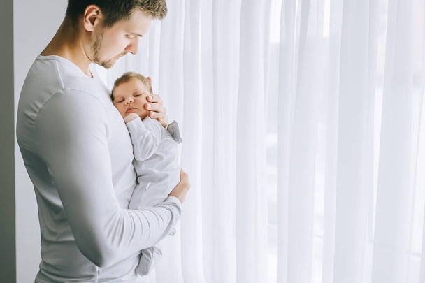 side view of handsome man carrying little baby boy in front of curtains at home - Foto, Imagen