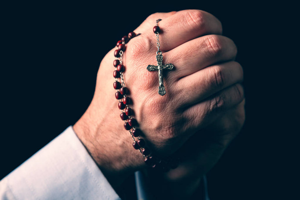 Мужские руки молятся держа четки с Иисусом Христом на кресте или распятие на черном фоне. Зрелый человек с христианской католической религией
 - Фото, изображение