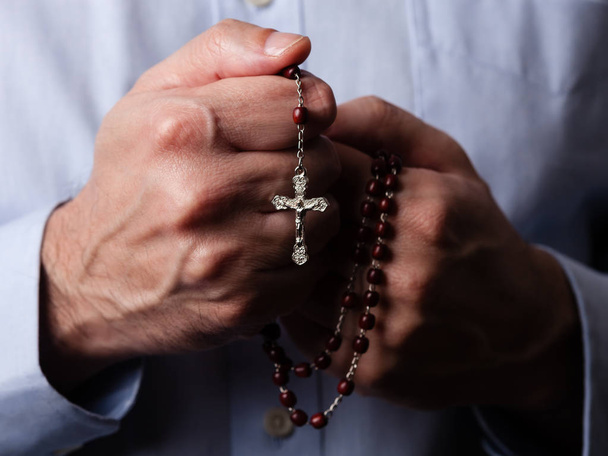 Male hands praying holding a rosary with Jesus Christ in the cross or Crucifix on black background. Mature man with Christian Catholic religious faith - Photo, Image