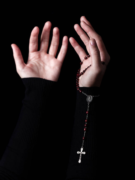 Female hands with arms outstretched praying and holding rosary with cross or Crucifix. Black background. Woman with Christian Catholic religious faith - 写真・画像