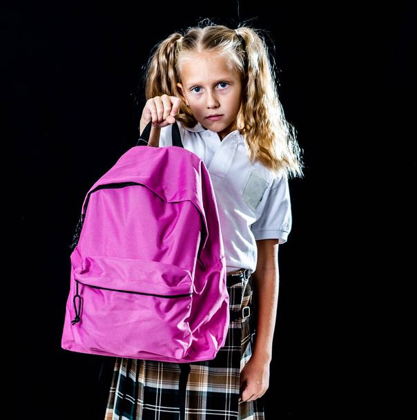 unhappy school girl holding a big schoolbag full of books and homework on black background. learning difficulties homework and education concept. - Photo, Image