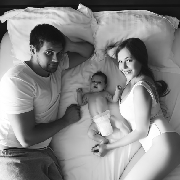μεγάλη γωνία θέα ευτυχισμένη οικογένεια στην τοποθέτηση με αξιολάτρευτο μικρό γιο το μωρό στο κρεβάτι στο σπίτι, μαύρο και άσπρο - Φωτογραφία, εικόνα