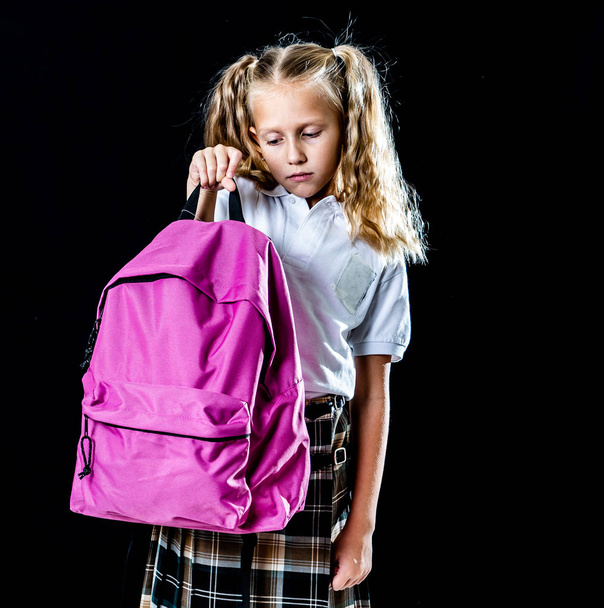 Sweet little girl in uniform carrying heavy big pink backpack or school bag on black background. School education concept - 写真・画像