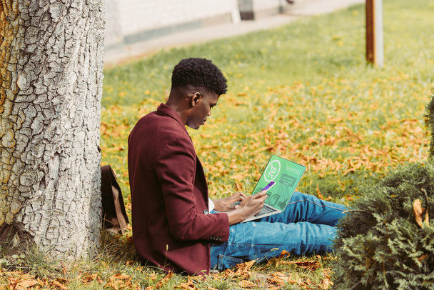 hombre afroamericano de compras en línea con el ordenador portátil y el teléfono inteligente mientras está sentado en la hierba en el parque
  - Foto, Imagen