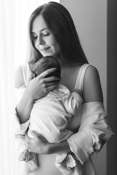 Schwarz-Weiß-Bild einer Mutter, die ihren kleinen Jungen zu Hause vor Vorhängen trägt - Foto, Bild