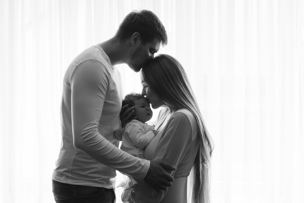black and white picture of man kissing wife while she kissing their little baby boy in front of curtains at home - Foto, Imagen