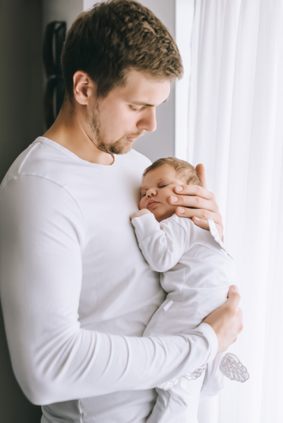 handsome father carrying little baby boy in front of curtains at home - Фото, изображение