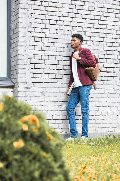 estudiante afroamericano con documentos y mochila de cuero de pie cerca de la pared de ladrillo blanco
 - Foto, Imagen