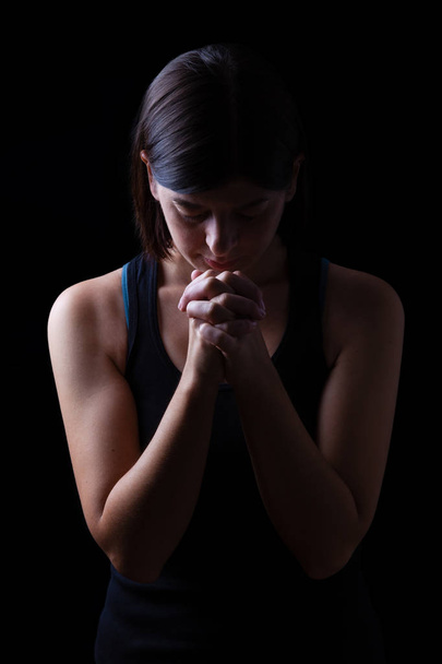 Faithful athletic woman praying, with hands folded in worship to god, head down and eyes closed in religious fervor, on low key black background. Concept for religion, faith, prayer and spirituality - Фото, изображение