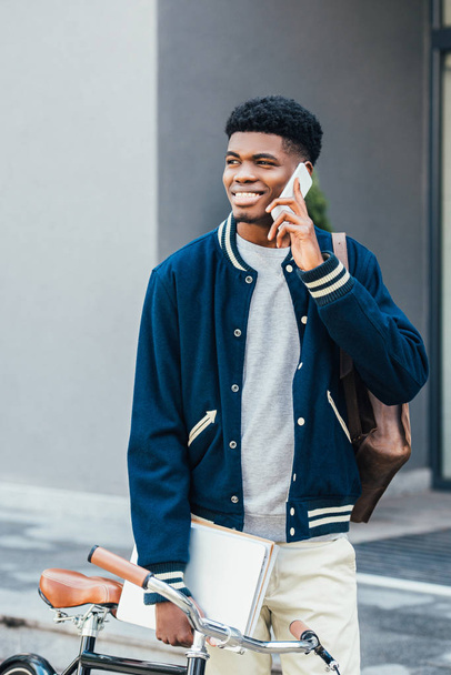 bell'uomo afroamericano sorridente con documenti che parlano su smartphone vicino alla bici
 - Foto, immagini
